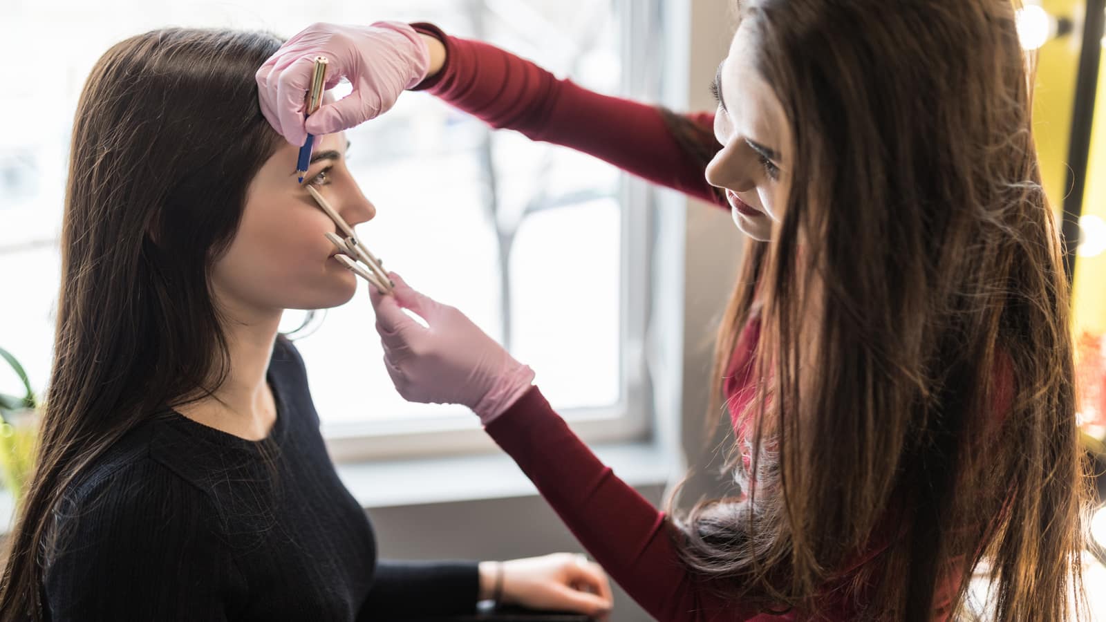 A brow technician in pink gloves measuring a client’s brow line with a pencil against a bright window