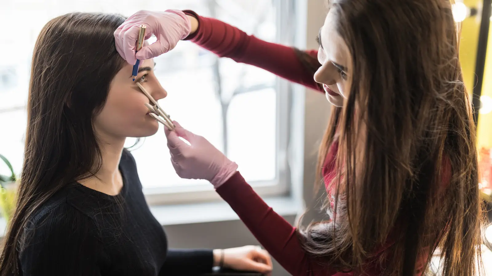 A brow technician in pink gloves measuring a client’s brow line with a pencil against a bright window