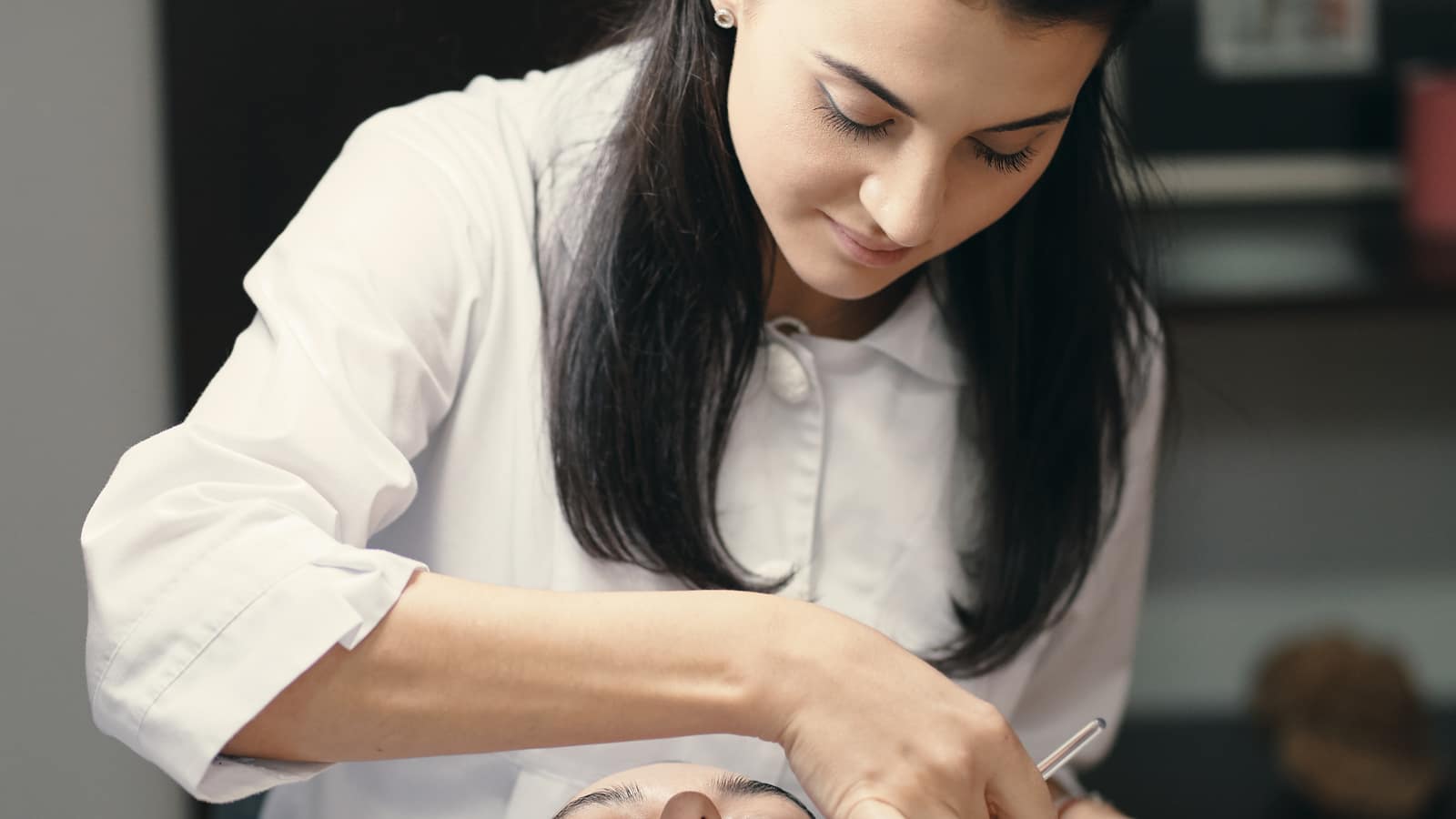 An esthetician in a white coat leaning over a client during a facial treatment