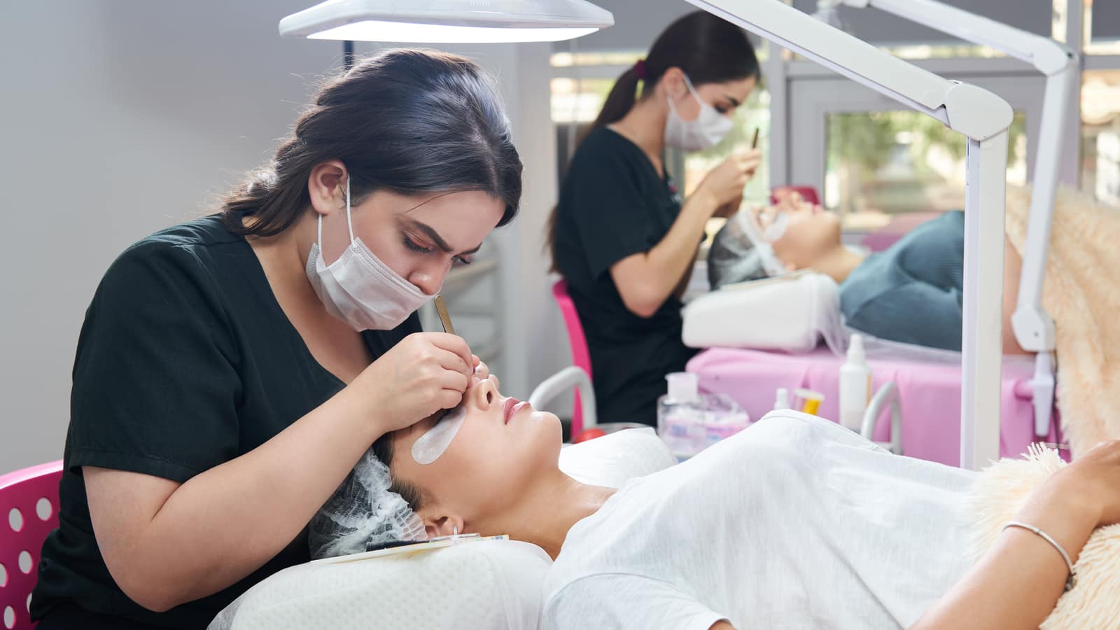 Two lash technicians working on clients at adjacent stations in a busy salon