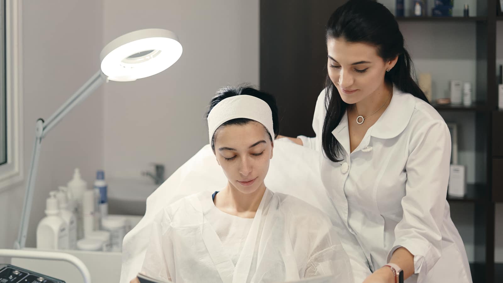 An esthetician in a white coat going through a treatment plan with a gowned client