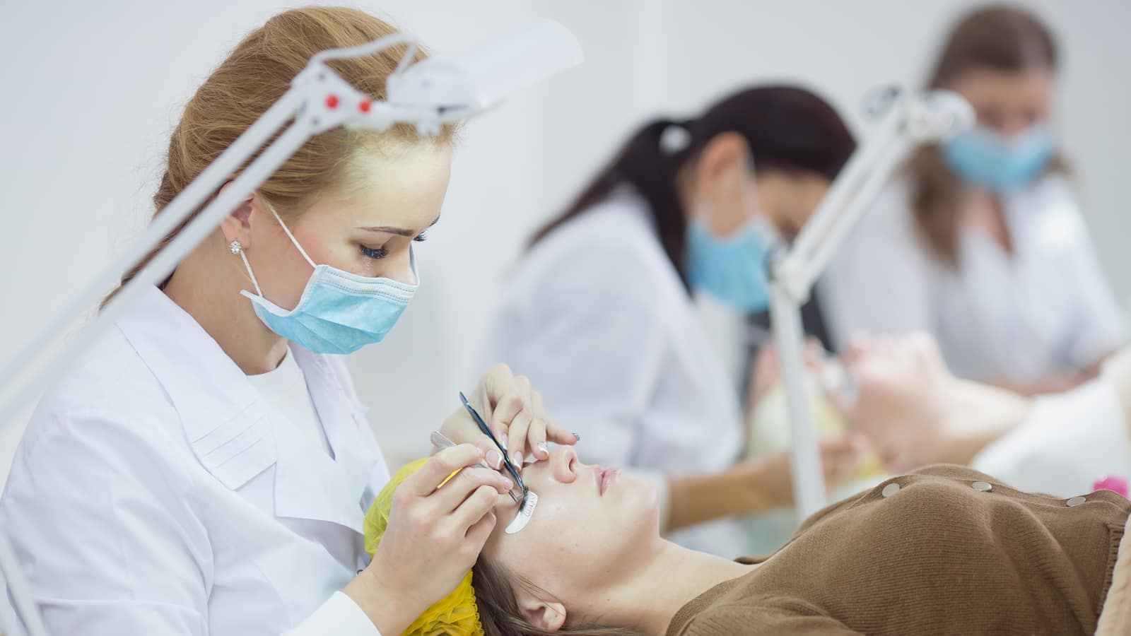 A lash technician in a mask applying extensions while two classmates work at tables behind her