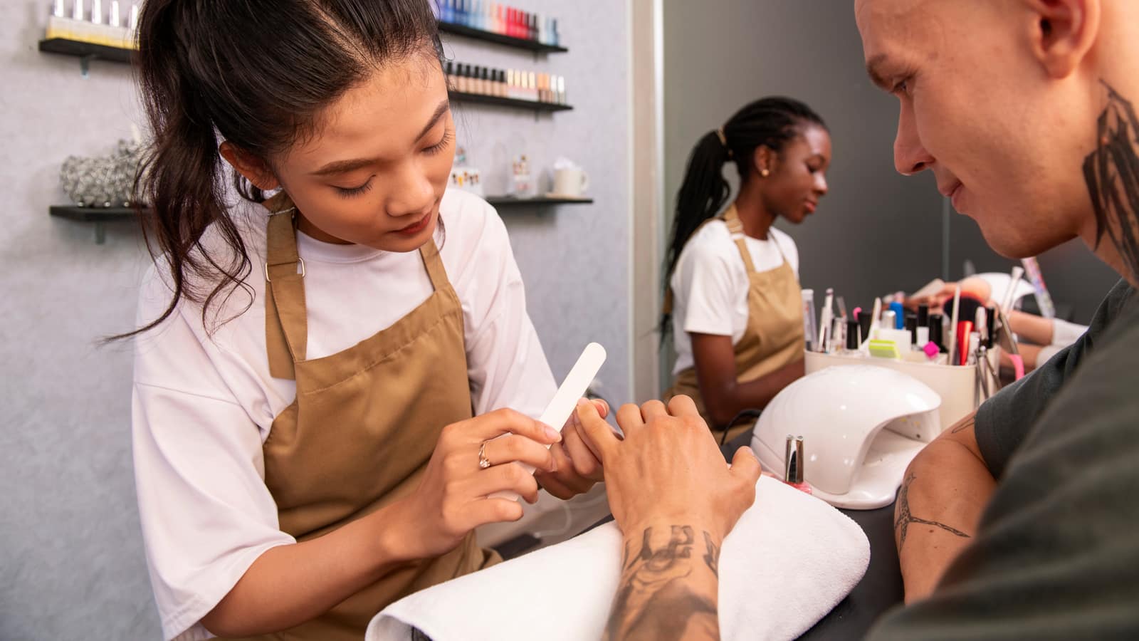 A nail technician filing a male client’s nails while a colleague works at the next station