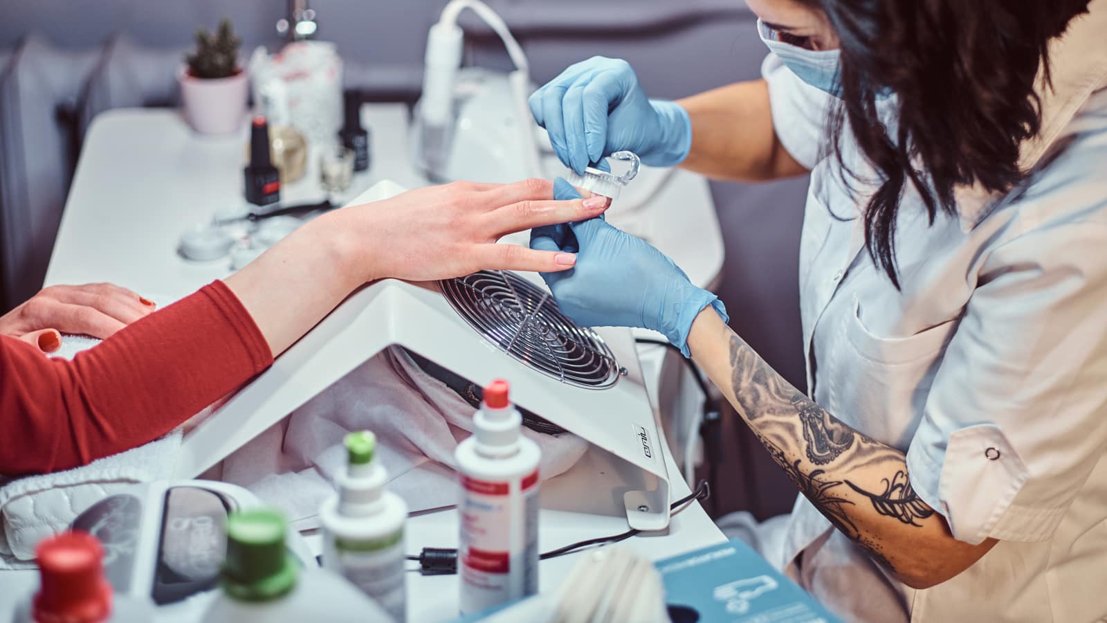 A masked nail technician buffing a client’s nails at a station with a dust extractor