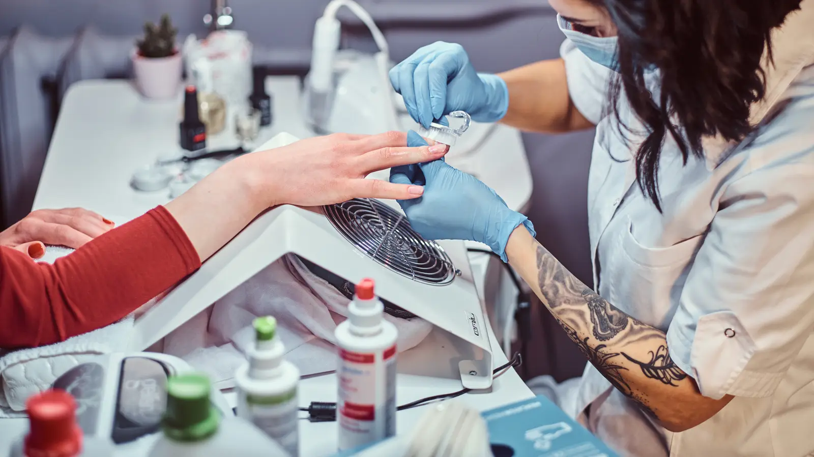 A masked nail technician buffing a client’s nails at a station with a dust extractor