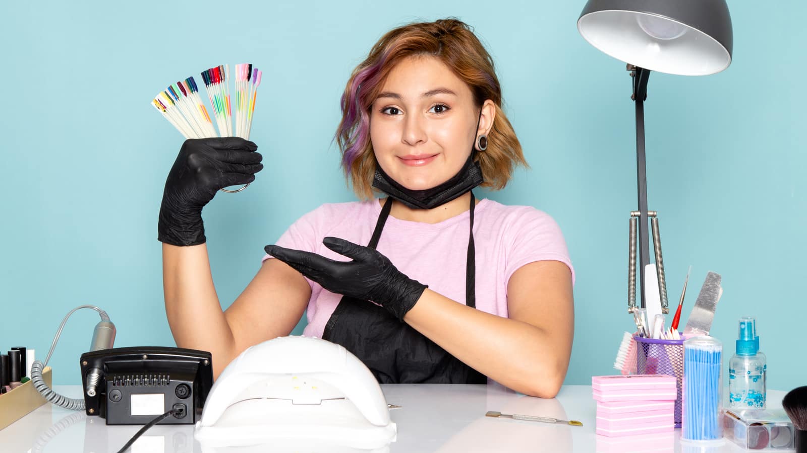 A nail technician in black gloves holding up a fan of colour sample sticks at a tidy work station
