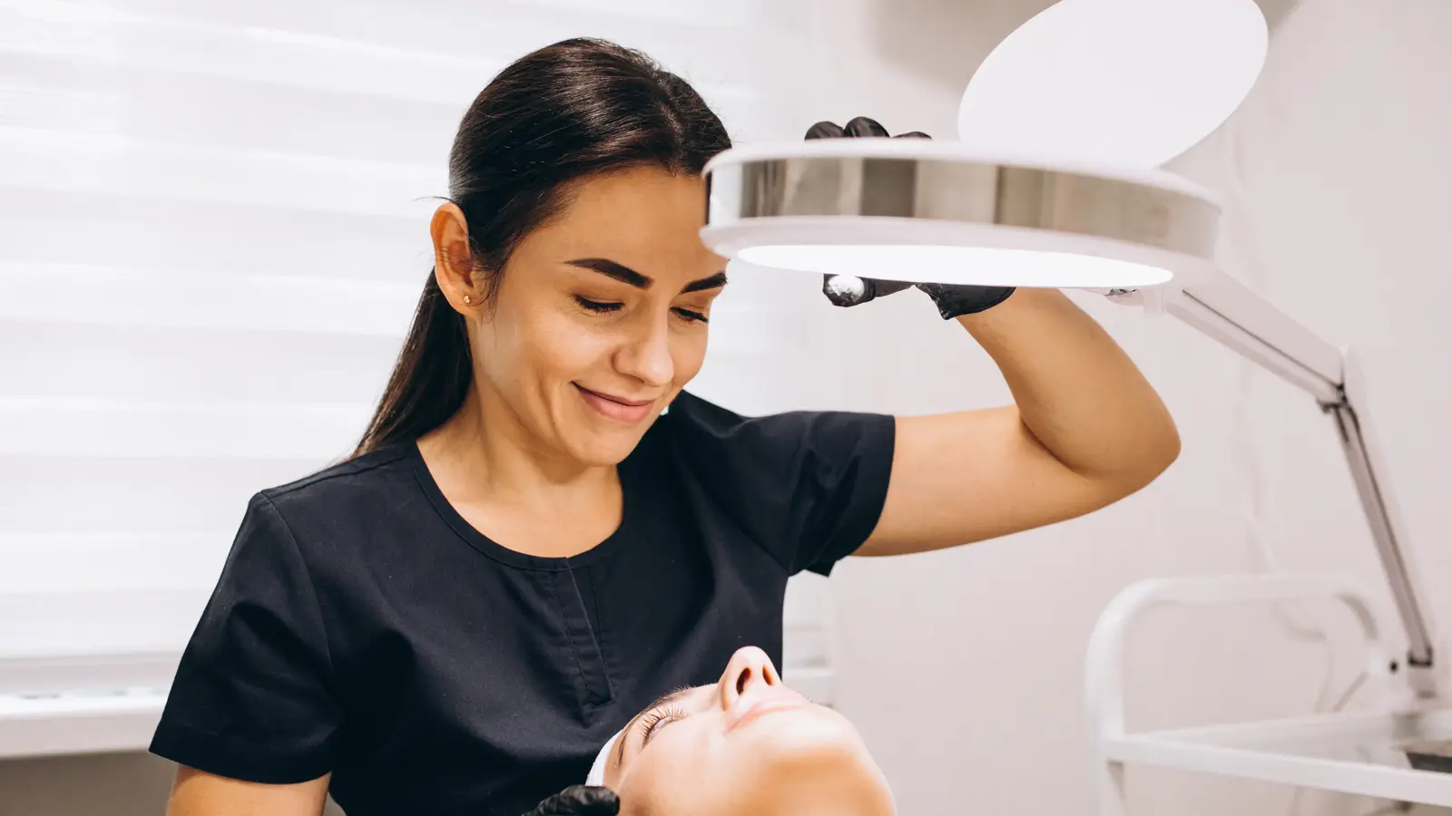 An esthetician in a black uniform and gloves angling a magnifying lamp over a client lying back on a treatment bed