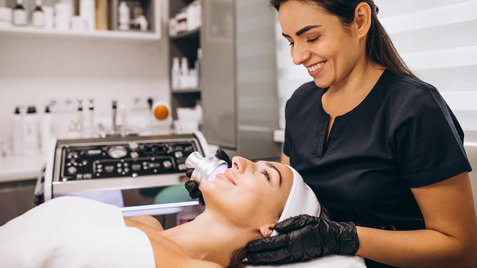An esthetician in black gloves running a handheld device over a client’s face in a treatment room lined with product shelves