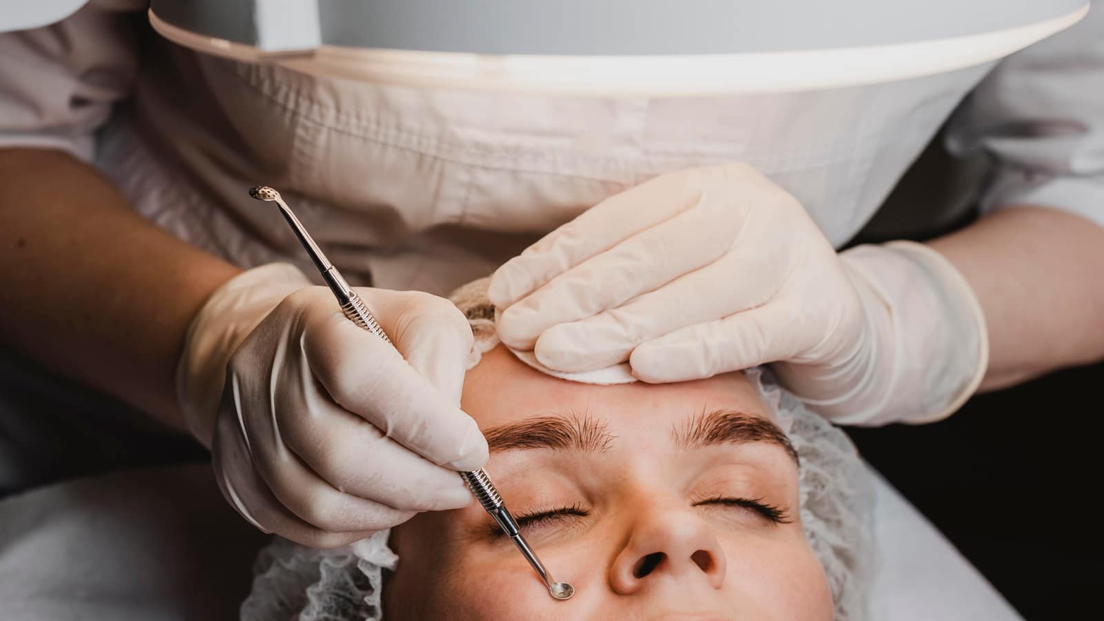 A gloved esthetician working on a client’s brow with extraction tools under a magnifying lamp
